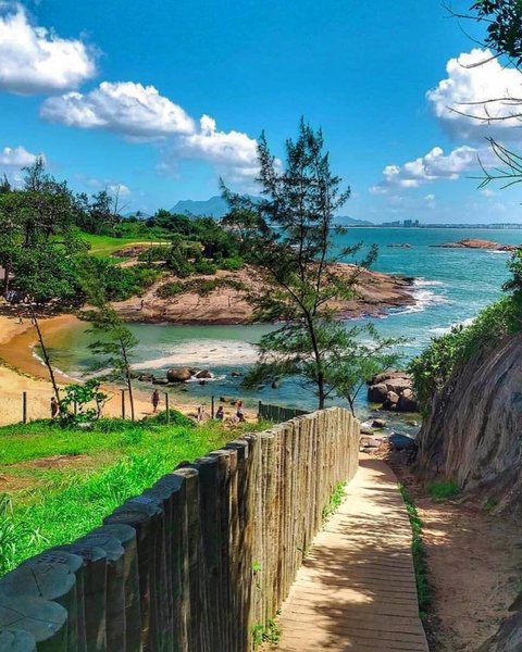 Caminho de madeira ao lado de uma cerca de troncos, descendo até a Praia Secreta, uma pequena praia de águas verdes entre pedras e árvores.